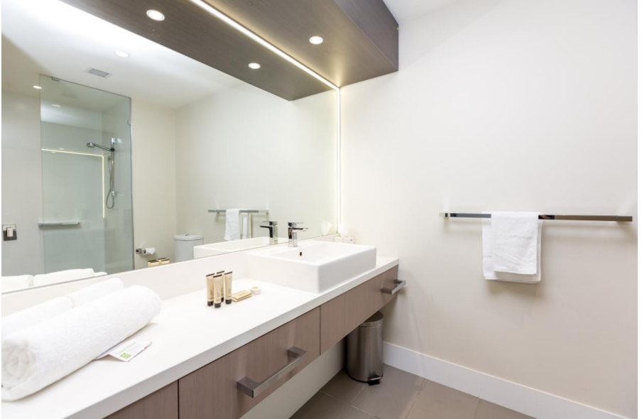 Redlands Rugby Union Club Modern bathroom with a large mirror, sink, white countertop, rolled towels, toiletries, wall-mounted towel rack with a white towel, and a glass-enclosed shower in the background.