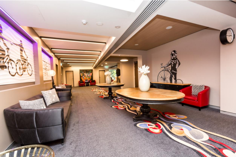 Redlands Rugby Union Club A modern lounge with black and red sofas, decorative tables, wall art featuring bicycles, a wall clock, and patterned carpet under ceiling lights.
