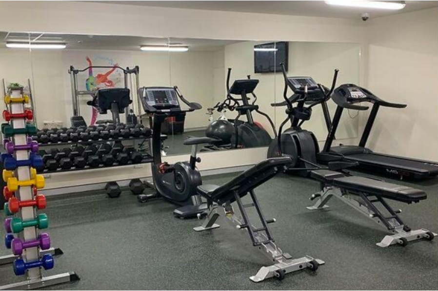 Redlands Rugby Union Club A gym room with free weights, two benches, a stationary bike, an elliptical machine, a treadmill, and a large mirror on the wall.