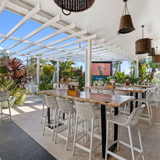 Redlands Rugby Union Club Open-air restaurant patio with wooden tables, white chairs, hanging wicker lamps, and lush green plants surrounding the seating area in daylight.