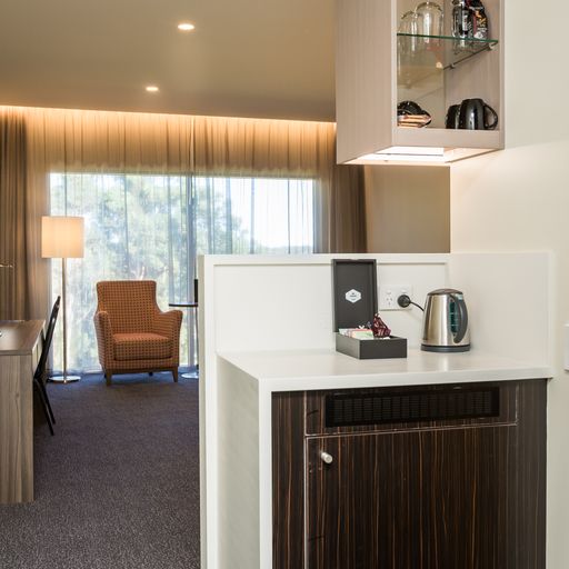 Redlands Rugby Union Club A hotel room features a seating area with an armchair by a window, a desk with a lamp, and a kitchenette with a kettle and cups on a shelf.