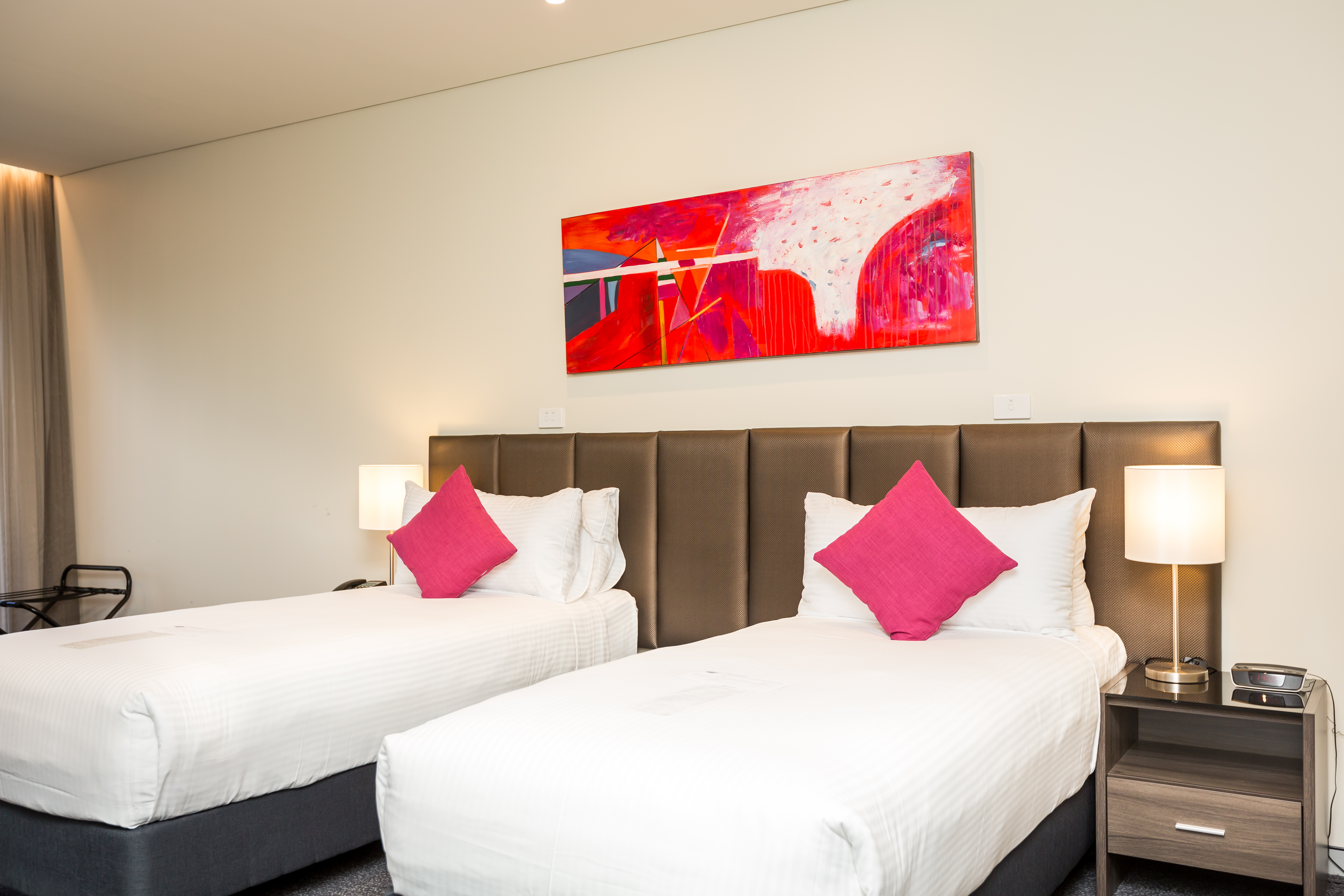 Redlands Rugby Union Club Two single beds with white linens and pink pillows, a colorful abstract painting above the headboard, bedside tables with lamps, and neutral-colored walls in a modern hotel room.