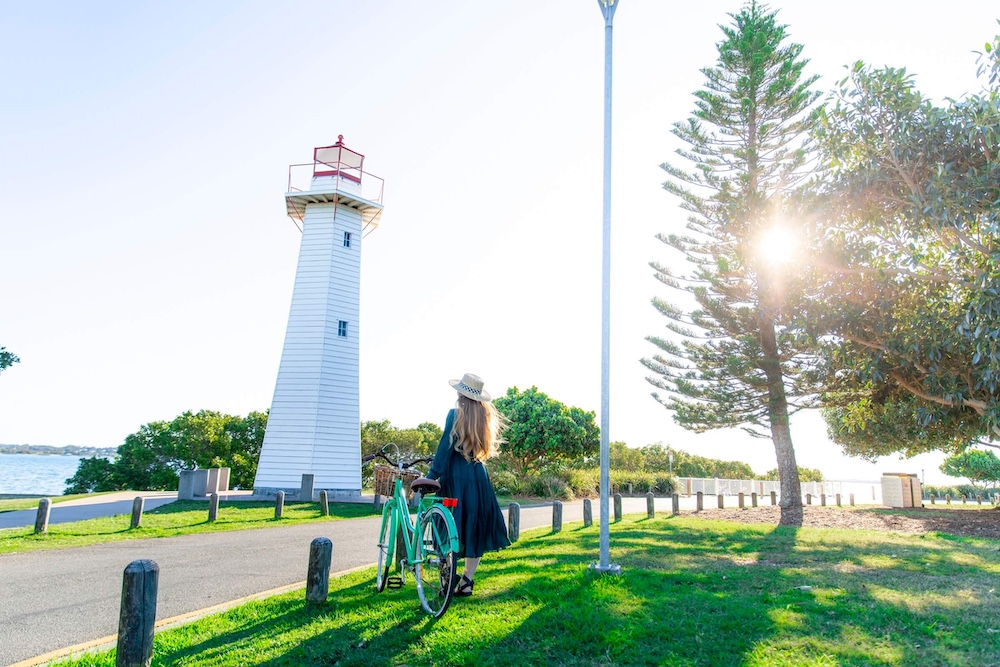 Redlands Rugby Union Club A person with a hat stands next to a green bicycle, looking at a white lighthouse near water, surrounded by trees on a sunny day.