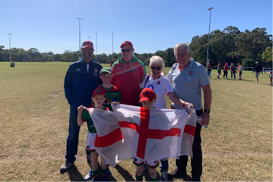 Redlands Rugby Union Club A group of adults and children stand on a sports field, smiling and holding a large England flag, with trees and other people in the background.
