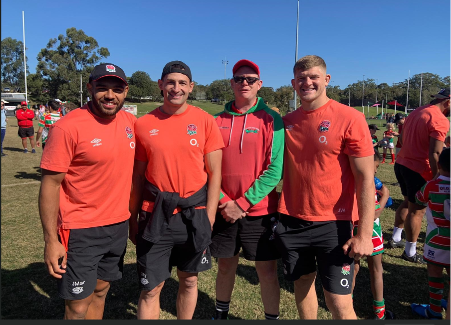 Redlands Rugby Union Club Four men stand outdoors on a grassy field, three wearing matching orange sports shirts and shorts, and one in a green and red jacket. Other people and sports activities are visible in the background.