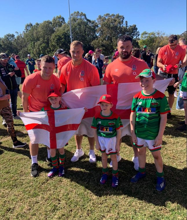 Redlands Rugby Union Club Three men in matching orange shirts stand behind three children wearing green rugby kits, holding England flags, on a grassy field with other people in the background.