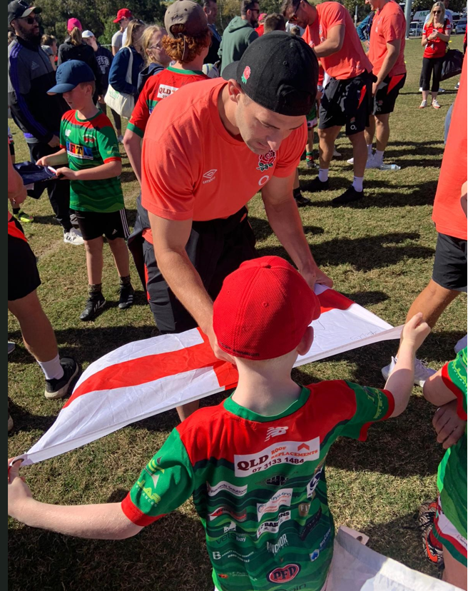 Redlands Rugby Union Club A man in a red sports shirt signs an England flag held by a child wearing a red cap and green sports jersey at an outdoor event.