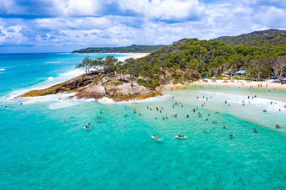Redlands Rugby Union Club Aerial view of a turquoise beach with swimmers and surfers in the water, trees and hills in the background, and partly cloudy skies overhead.
