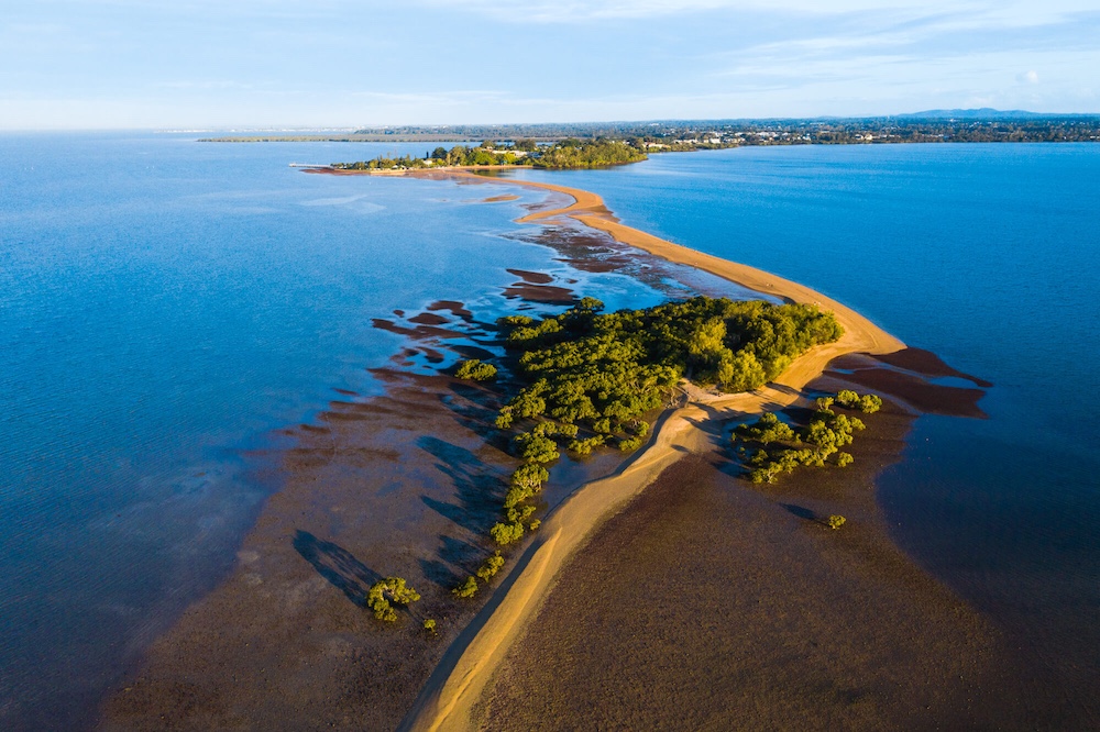 Redlands Rugby Union Club Aerial view of a narrow sandbar extending into the ocean, with patches of vegetation and distant buildings on the coastline under a clear sky.